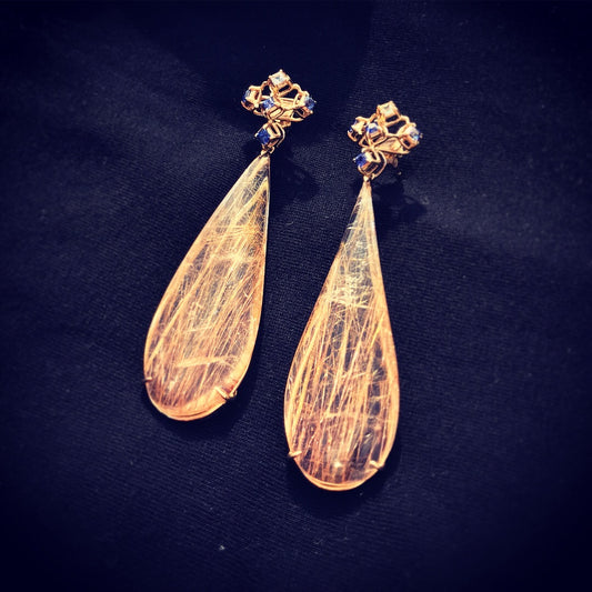 A pair of 14 karat yellow gold dangling earrings with Blue and White Sapphires. Its top part consists of five square cut, top is white and the others are blue sapphires. The bottom dangling part is a tear drop shaped Rutilated Quartz. They are lying down on the black cloth like stargazing from the ground.