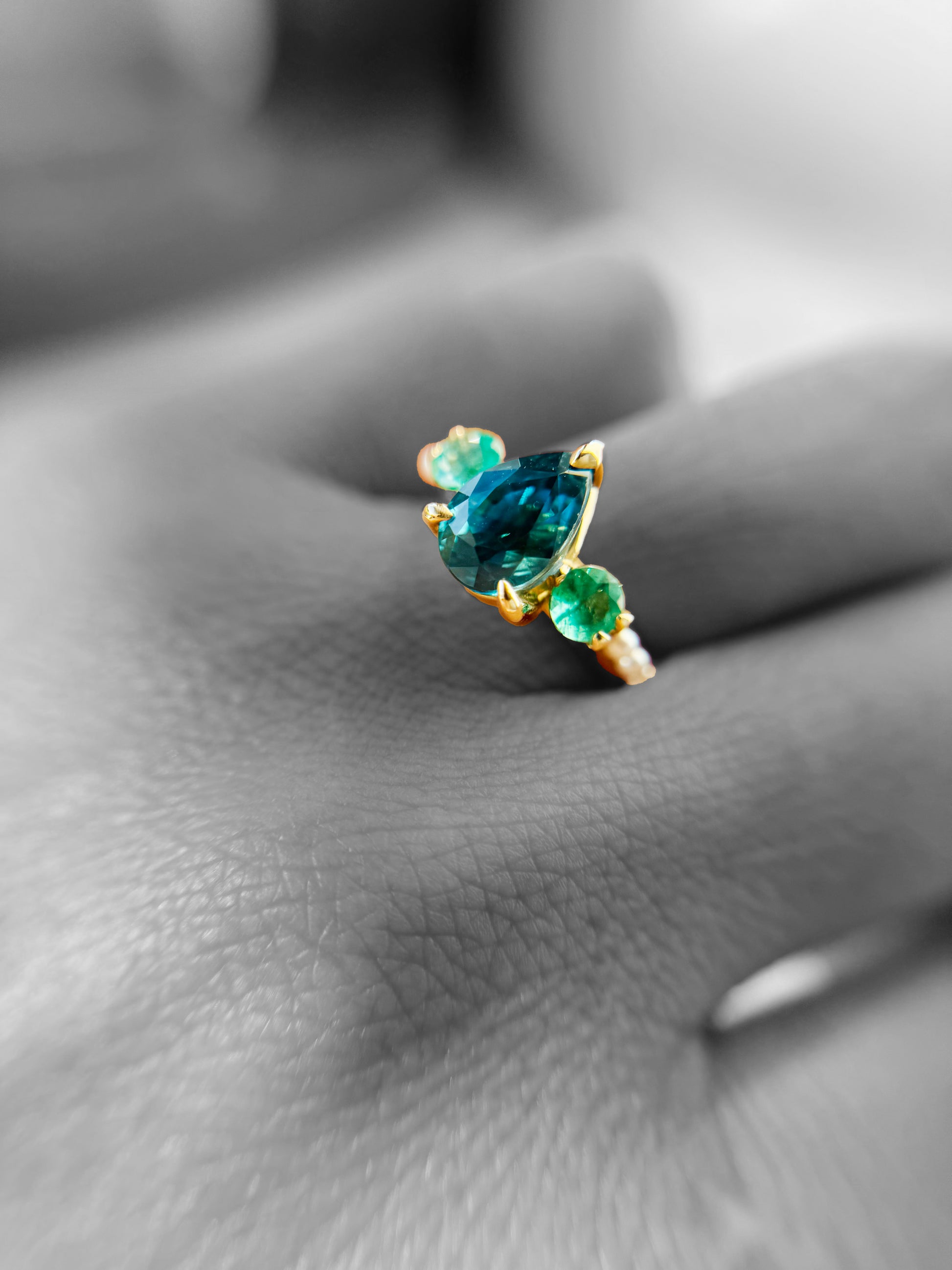 Gold ring is worn one hand. Background is monotone. Pear shaped blue Zircon is prong set in between two side round cut Emeralds. Three tiny white pearls are lined up on its shoulders. 