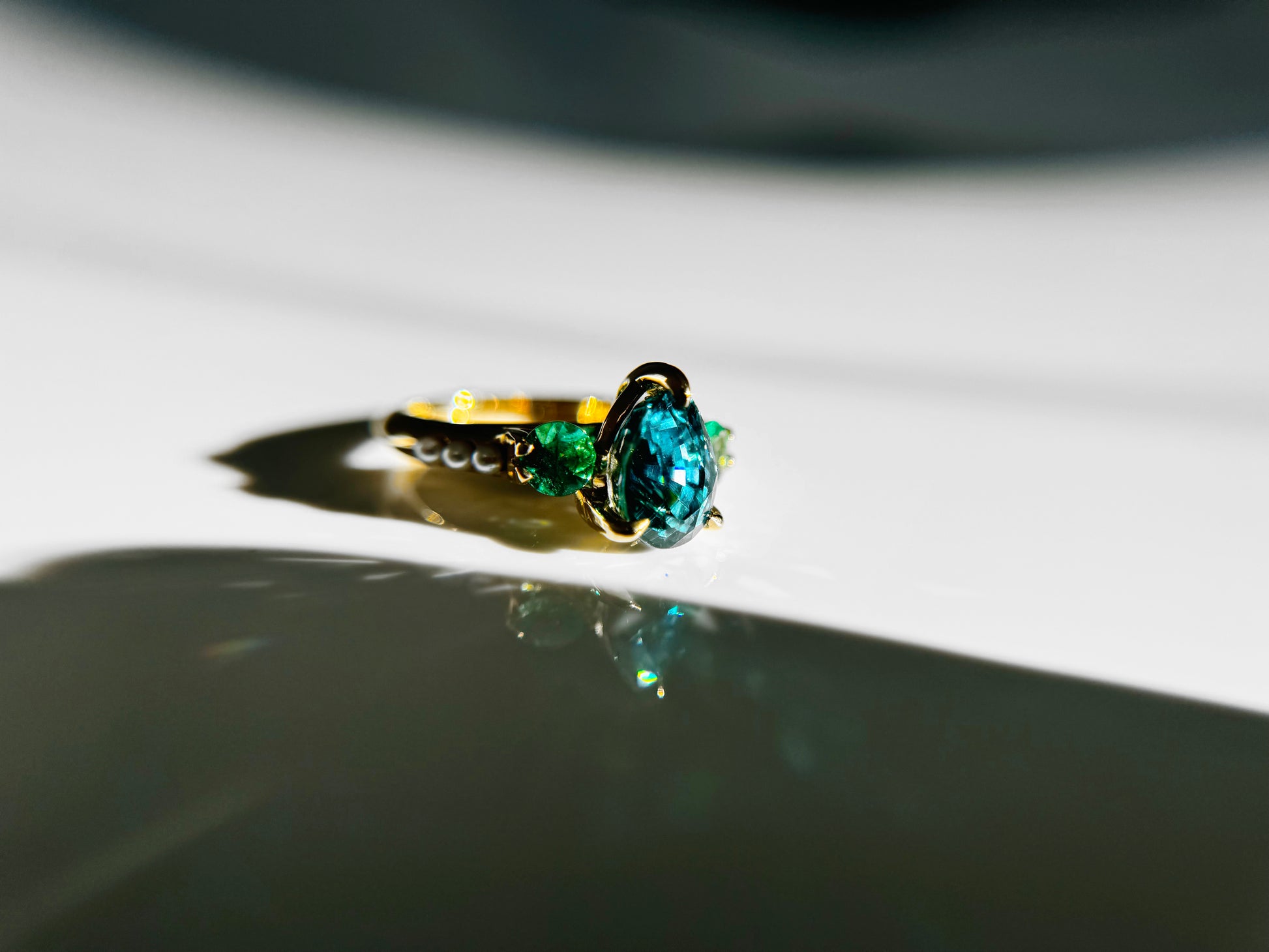 The side view of a yellow gold ring. Pear shaped blue Zircon is prong set in between two side round cut Emeralds. Three tiny white pearls are lined up on its shoulders. 