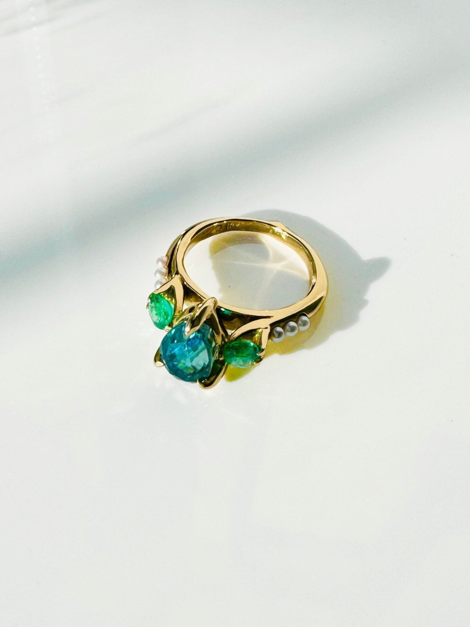 Pear shaped blue Zircon is prong set in between two side round cut Emeralds. Three tiny white pearls are lined up on its shoulders. This yellow gold ring is laying down on a white table. 