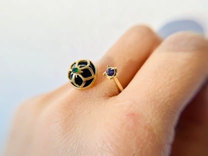 Wearing a 14 karat and 18 karat yellow gold bypass ring featuring a cutout balloon flower design with a tiny Tsavorite garnet on the top of dome shaped onyx. There is a 3mm purple sapphire on the other ring end